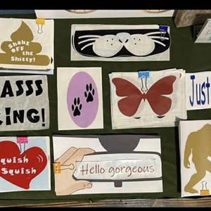 May include: A collection of handmade signs with various designs and text. The signs include phrases like "LORD", "YASSS KING!", and "Hello gorgeous". Other signs feature a butterfly, paw prints, and a Bigfoot silhouette. The signs are attached with clips.