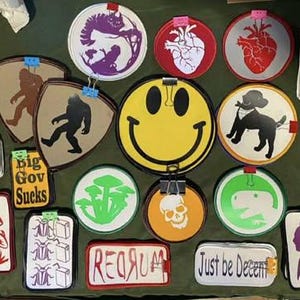 May include: A collection of various round and rectangular stickers and patches with diverse designs. The designs include a smiley face, a heart, a dog, a skull, mushrooms, and the text "HUMAN", "GOD > GOV", "Big Gov Sucks", "REORUM", and "Just be Decent".