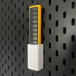 May include: A yellow and black pegboard tool holder with a white base. The holder has a rectangular shape with a series of black, grooved sections. The base is a solid white block. The background is a black pegboard.