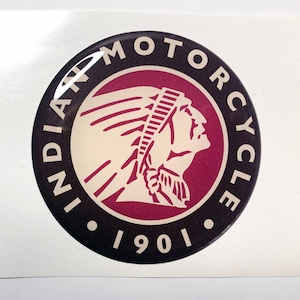 May include: A circular badge featuring the Indian Motorcycle logo. The design includes a profile of a Native American in a headdress, set within a maroon circle, with the text "INDIAN MOTORCYCLE" and the year "1901".