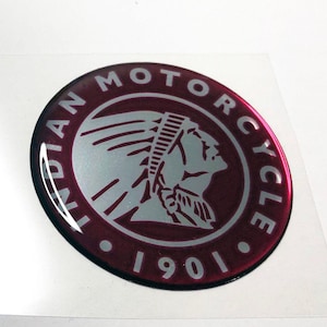 Puede incluir: Una pegatina circular con las palabras "INDIAN MOTORCYCLE" en letras blancas alrededor del borde. El centro presenta un perfil plateado de un jefe nativo americano. El año "1901" está en la parte inferior. La pegatina tiene un fondo granate brillante.