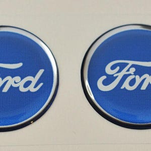 Puede incluir: Dos emblemas circulares de Ford con fondo azul y letras blancas. Los emblemas tienen un borde plateado y se muestran sobre un fondo blanco. El logotipo de Ford es un diseño automotriz clásico.