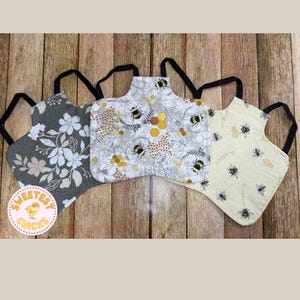 Puede incluir: Tres pequeños delantales con correas negras, cada uno con un patrón único. Uno es gris con flores blancas, otro tiene abejas y panales sobre un fondo gris, y el tercero es amarillo claro con abejas negras. Los delantales están sobre una superficie de madera.
