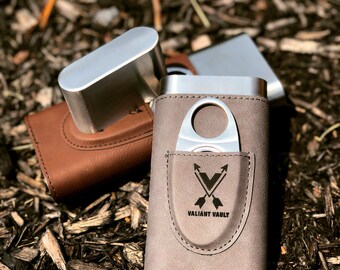 Puede incluir: Un estuche de cuero marrón para cigarros con un cortador de cigarros de metal plateado y un porta cigarros de metal plateado. El estuche tiene las palabras "Valiant Vaults" grabadas.