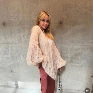 Peut inclure: Un gilet beige en tricot ajouré avec des manches longues et amples et une fermeture boutonnée, associé à une jupe midi rouge foncé à petit motif géométrique blanc et un petit sac à main avec une bandoulière en chaîne argentée.