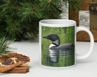 May include: White ceramic mug featuring a colourful image of a loon swimming in a lake with green reeds. The loon has black and white feathers and a red eye.