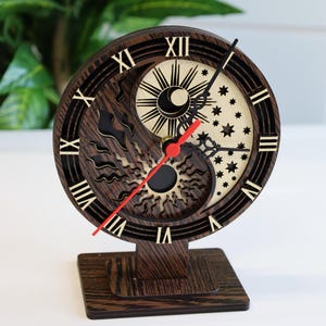 May include: A wooden clock with a yin yang design featuring a sun and moon. The clock face has Roman numerals and a red hour hand and black minute hand. The clock is on a wooden stand.