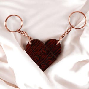 Puede incluir: Un llavero de madera en forma de coraz&oacute;n con un patr&oacute;n de veta de madera. El llavero est&aacute; unido a dos llaveros de color dorado.