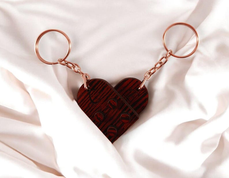 Puede incluir: Un llavero de madera en forma de coraz&oacute;n con un patr&oacute;n de veta de madera. El llavero est&aacute; unido a dos llaveros de color dorado.