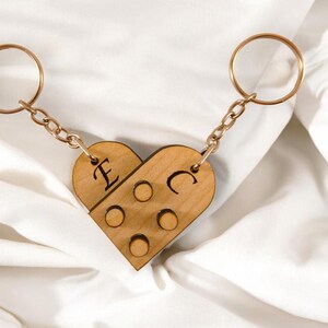 Puede incluir: Un llavero de madera con forma de coraz&oacute;n con las letras "E" y "C" grabadas. El llavero est&aacute; hecho de dos piezas que encajan, y tiene una cadena dorada y dos anillos.