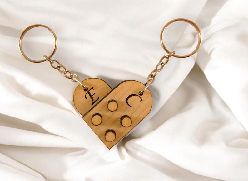 Puede incluir: Un llavero de madera con forma de coraz&oacute;n con las letras "E" y "C" grabadas. El llavero est&aacute; hecho de dos piezas que encajan, y tiene una cadena dorada y dos anillos.