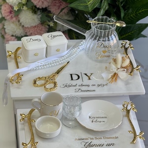 May include: A white marble tray with gold accents and the text "DY Kabul olmus en guzel Duamsin"  A white ceramic mug with a gold rim and the letter "V" on it. A white ceramic bowl with a gold rim. A clear glass with a gold rim. A white ceramic plate with the text "Kıyamam bilirsin" and a gold rim. A white marble tray with gold accents and the text "Annem'in Son Damadı Yunus un Damat Kahvesi".