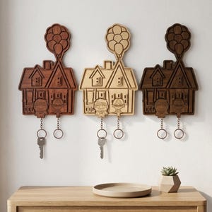 Puede incluir: Tres llaveros de madera con forma de casas, cada uno con un racimo de globos y figuras de personajes. Los llaveros son en tonos marrones y beige, con llaves colgando debajo. Están montados en una pared blanca.