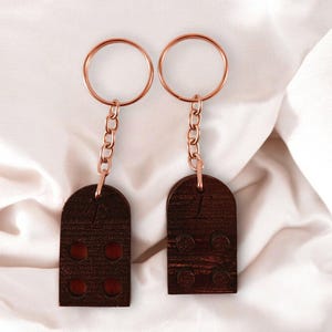 Puede incluir: Dos llaveros de madera marr&oacute;n con anillos y cadenas de metal dorado. Cada llavero tiene un n&uacute;mero diferente grabado. El llavero de la izquierda tiene el n&uacute;mero "1" grabado. El llavero de la derecha tiene el n&uacute;mero "2" grabado.