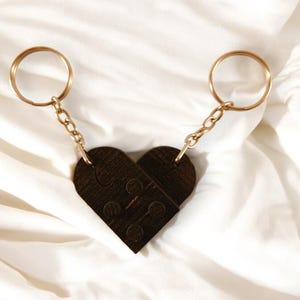 Puede incluir: Dos llaveros dorados con un dije de madera marr&oacute;n en forma de coraz&oacute;n. El dije est&aacute; hecho de dos piezas que encajan como un rompecabezas.