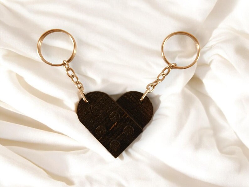 Puede incluir: Dos llaveros dorados con un dije de madera marr&oacute;n en forma de coraz&oacute;n. El dije est&aacute; hecho de dos piezas que encajan como un rompecabezas.