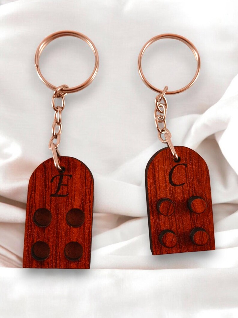 Puede incluir: Dos llaveros de madera con un anillo y una cadena de metal dorado. Los llaveros tienen forma de casita peque&ntilde;a con una letra 'E' y 'C' grabadas en la parte delantera. Cada llavero tiene cuatro peque&ntilde;os c&iacute;rculos en la parte delantera.