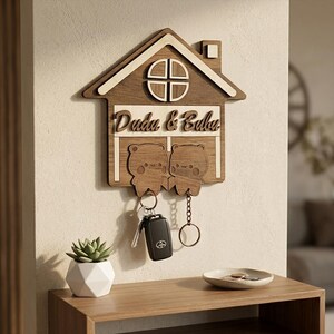 May include: Wooden house-shaped key holder with the names "Dudu & Bubu" engraved. Features two bear-shaped key hangers and a key with a car remote attached. The design is in shades of brown and white, mounted on a wall.