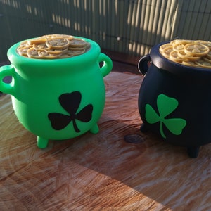 Könnte beinhalten: Zwei dekorative Töpfe mit Goldmünzen, einer grün und einer schwarz, stehen auf einer Holzoberfläche. Jeder Topf ist mit goldfarbenen Münzen gefüllt und hat ein Kleeblatt-Design. Die Töpfe haben kleine Füße und Griffe.