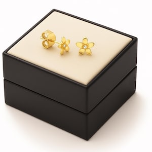 Könnte beinhalten: Goldfarbene Blüten-Ohrstecker mit einem kleinen Diamanten in der Mitte jeder Blüte. Die Ohrringe werden auf einer beigen Oberfläche in einer schwarzen Schmuckschatulle präsentiert.