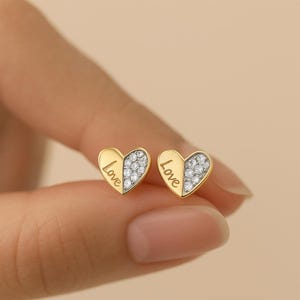 May include: A pair of gold-coloured heart-shaped stud earrings. One half of each heart is covered in small, clear crystals, while the other half has the word "Love" engraved. The earrings are held between fingers.