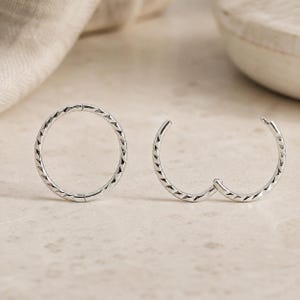 May include: A pair of silver hoop earrings featuring a textured, faceted surface. One earring is shown as a complete circular hoop, while the other is displayed as an open, hinged hoop, highlighting the metallic finish and the geometric pattern.