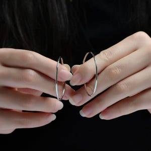 May include: A pair of silver hoop earrings. The earrings are held up by two hands against a black background. The hoops are thin and circular, with a polished, reflective surface.