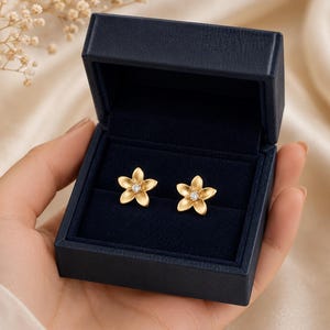 May include: A pair of gold flower-shaped stud earrings with a central clear stone, presented inside a dark navy blue rectangular jewellery box with a velvet-lined interior. The earrings feature five petals each with a matte finish and a central setting.