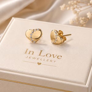 May include: A pair of gold heart-shaped stud earrings featuring a curved line of small clear stones set across the centre. The earrings are displayed on a cream-coloured box with the text In Love JEWELLERY printed in gold lettering on the lid.