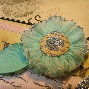 Könnte beinhalten: Eine hellblaue Papierblume mit silbernen Glitzerakzenten und einer braunen Mitte mit silbernem Glitzer. Die Blume befindet sich auf einem braunen Hintergrund mit einem rosa Band.