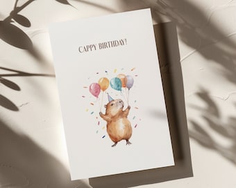 May include: A birthday card featuring a watercolour illustration of a guinea pig wearing a party hat and holding onto colourful balloons. The card reads "Cappy Birthday!"