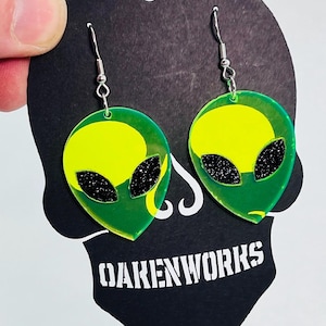 May include: A pair of green and yellow alien face earrings with black glitter eyes. The earrings are on a black card with the text "DAKENWORKS" printed in white.