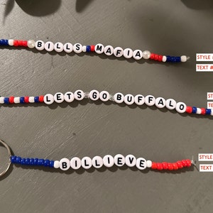 Könnte beinhalten: Drei Schlüsselanhänger mit Perlen und silbernen Verschlüssen. Der erste Schlüsselanhänger trägt die Aufschrift "BILLS MAFIA" in weißen Perlen mit roten, weißen und blauen Akzenten. Der zweite lautet "LETS GO BUFFALO". Der dritte lautet "BILLIEVE".