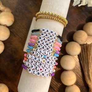 May include: A stack of colorful beaded bracelets with words like "Mama", "Belly", "Boy", "Girl", "Mom", "Everest", "Love", and "Penny" spelled out on them.