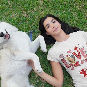 May include: A white t-shirt with a pink and red graphic design of the word "LOVE" surrounded by hearts and an angel with an arrow. A woman is wearing the shirt and is lying on the grass with a white and grey dog.