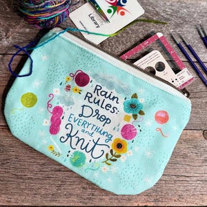 May include: A light blue zippered pouch featuring the text "Rain Rules: Drop EVERYTHING and Knit" surrounded by colorful yarn balls and flowers. Included are a ball of variegated yarn, scissors, a library card, a needle gauge, and four knitting needles.