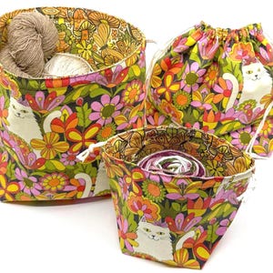 May include: Three fabric storage containers with a colorful floral and cat print. The containers are in shades of green, orange, yellow, and pink. One container is a drawstring bag, and the other two are open-top baskets. Yarn is visible inside the baskets.