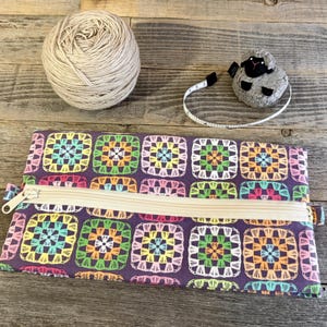 May include: A rectangular zippered pouch featuring a repeating pattern of colorful granny square motifs on a dark purple background, accompanied by a beige ball of yarn and a sheep-shaped measuring tape with a white tape measure.