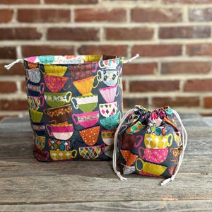 May include: Two fabric drawstring bags. The larger bag is a tall cylinder, and the smaller bag is a pouch. Both are patterned with colorful teacups on a dark gray background. The bags have white drawstrings and are sitting on a wooden surface.