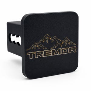 May include: A black square trailer hitch cover featuring a textured finish. The front face displays an engraved mountain range graphic positioned above the word TREMOR in bold, capitalized lettering. The side shows a square receiver tube.