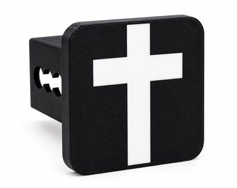 Peut inclure: Un cache-attelage de remorque noir présentant une face carrée avec un symbole de croix blanche centré sur le devant. Le cache-attelage possède un tube de montage carré s'étendant à l'arrière avec trois trous circulaires visibles sur le côté.