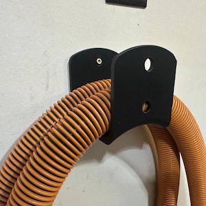 Könnte beinhalten: Schwarze Wandhalterung mit zwei daran befestigten orangefarbenen Spiralschläuchen. Die Halterung hat zwei Löcher für Schrauben.