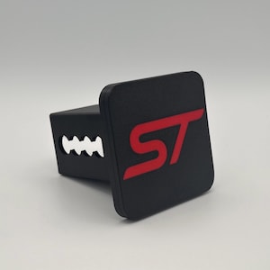 Peut inclure: Cache-attelage noir avec le logo "ST" rouge. Le cache est carré avec des bords arrondis et une base rectangulaire avec des découpes. Le logo est dans une police audacieuse et stylisée.