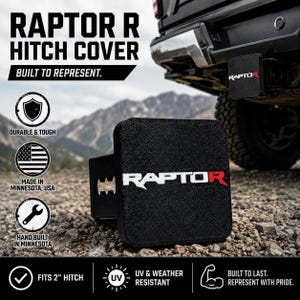 May include: A black square hitch cover featuring the white and red text RAPTOR R. The accessory is designed to fit a 2-inch hitch receiver. It has a textured finish and is UV and weather resistant, built to be durable and tough for vehicle use.