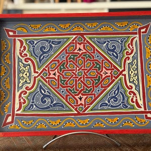 Könnte beinhalten: Ein Holztablett mit rotem Rand und einem blauen bemalten Design. Das Design zeigt komplizierte geometrische Muster in Rot, Gelb, Grün und Blau.