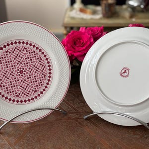 Könnte beinhalten: Zwei weiße Keramikteller mit einem dekorativen rosa und weißen Muster auf einem Teller und einem rosa Logo auf dem anderen. Die Teller stehen auf Metallständern.