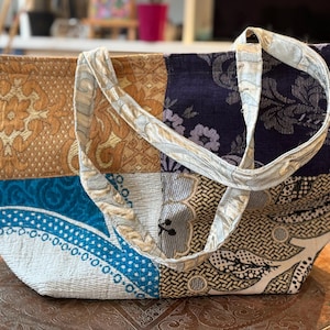 Peut inclure: Un sac fourre-tout patchwork avec divers tissus à motifs dans les tons or, bleu et marine. Le sac a une forme rectangulaire avec une large ouverture et deux anses. Le sac est posé sur un plateau marron.
