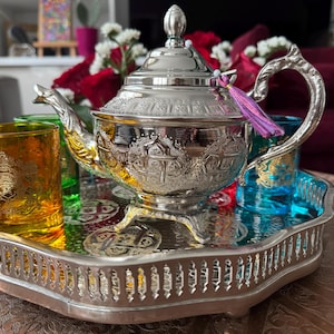 Peut inclure: Une théière en argent avec des motifs complexes est posée sur un plateau en argent avec quatre petites tasses en verre. Les tasses sont remplies de liquides colorés et la théière a un pompon violet.