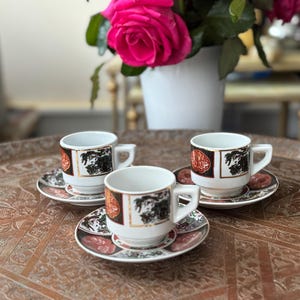 Puede incluir: Tres tazas de té de cerámica blanca con diseños florales negros y dorados en una bandeja marrón. Cada taza tiene un platillo a juego con un diseño floral rojo y dorado.