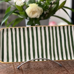 Könnte beinhalten: Ein rechteckiges Serviertablett in Weiß mit grünen Streifen und goldfarbenem Rand. Das Tablett steht auf einem Ständer.